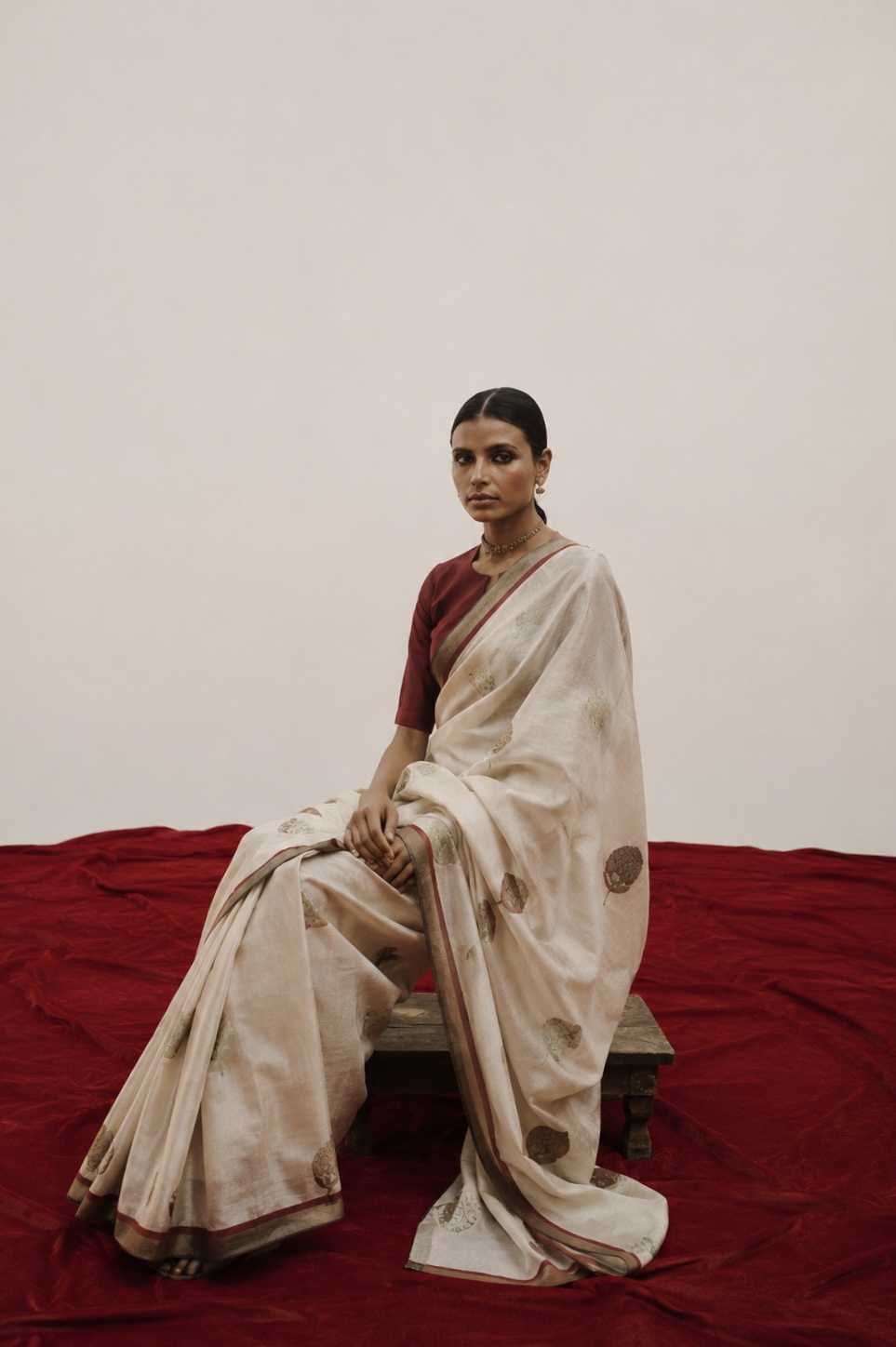 Woman in a cream saree seated on red velvet